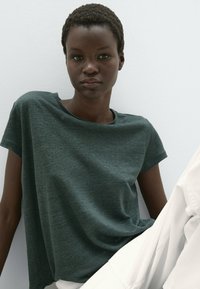 Donkergroene, korte mouwen, relaxte fit t-shirt van zachte stof. Kenmerkend is de ronde halslijn en subtiele textuur, gecombineerd met witte broek.