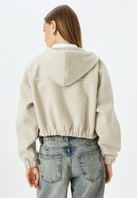 Zweifarbiger, hellgrauer Hoodie mit texturiertem Finish, ausgestattet mit einer Kapuze und einem elastischen Bund. Kombiniert mit hoch taillierten blauen Jeans.