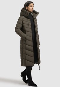 Lange, olivenfarvet pufferjakke med høj krave, quiltet design, lynlås foran og sidelommer. Sammen med sorte højhælede støvler.