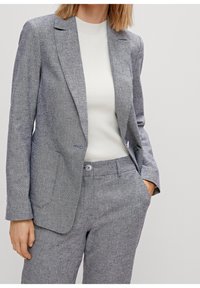 Blazer grigio su misura realizzato in tessuto testurizzato, con due tasche frontali, rever a notched e una vestibilità slim, abbinato a un top bianco a coste.