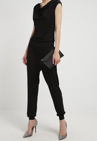 Top negro drapeado combinado con pantalones negros sueltos. La modelo sostiene un bolso de mano tejido negro y lleva zapatos de tacón alto plateados con acabado metálico.
