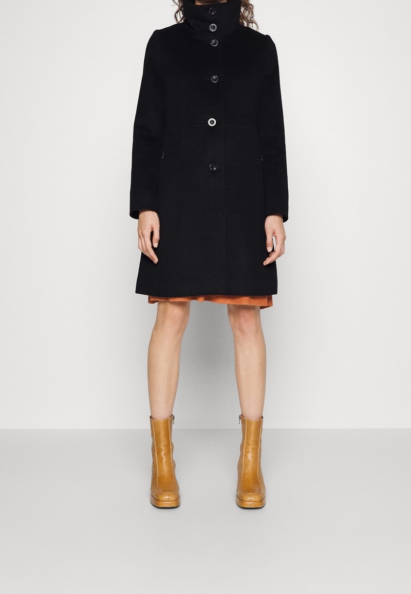 Femme portant un manteau noir boutonné, une jupe couleur rouille et des bottines beige, debout devant un fond clair uni.