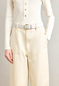 Kräftribbad långärmad topp i crème med knappdetalj, tillsammans med högmidjade, vida beige byxor och ett vitt bälte med en guldfärgad spänne.