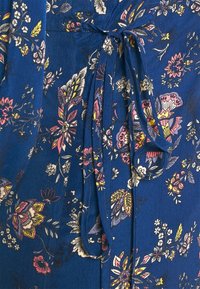 Tissu floral bleu présentant des motifs intricats en rose, jaune et blanc, avec un détail de taille nouée. Texture douce et design fluide.