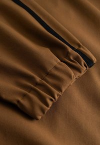 Gros plan sur des manches en tissu marron avec un détail de bande noire et des poignets élastiqués sur un vêtement.