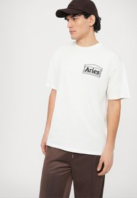 T-shirt en coton blanc avec un logo "Bélier" imprimé en noir sur la poitrine gauche, assorti d'un pantalon de sport marron et d'une casquette foncée.