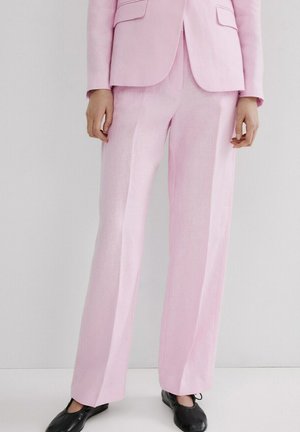 Persona vestida con pantalones y chaqueta rosa claro a medida, y bailarinas negras, de pie frente a un fondo claro y liso.