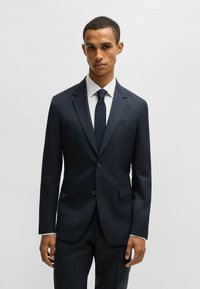 Completo blu a righe sottili con vestibilità su misura, due bottoni e rever a punta. Abbinato a una camicia bianca a righe e una cravatta scura.