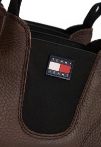 Bota de cuero texturizado marrón con panel elástico negro que presenta un logo rectangular de Tommy Jeans en blanco y rojo.