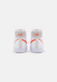 Un paio di sneaker alte bianche Nike con swoosh arancione e logo sul retro, viste da dietro su uno sfondo bianco.