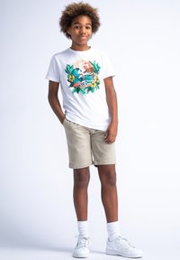 T-shirt bianco con una grafica tropicale colorata, abbinato a pantaloni corti beige. Sneakers bianche e calzini alla caviglia completano l'outfit.