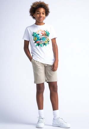 T-shirt blanche avec un graphique tropical coloré, associé à un short beige. Des baskets blanches et des chaussettes mi-mollet complètent la tenue.