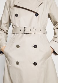 Beżowy płaszcz trencz o podwójnym zapięciu, z dużymi czarnymi guzikami, paskiem w talii i bocznymi kieszeniami, wykonany z gładkiego materiału.