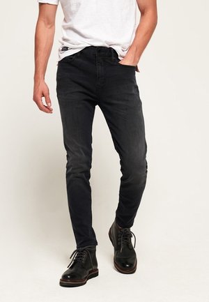 Man draagt zwarte skinny jeans, zwarte veterlaarzen en een wit T-shirt met korte mouwen, met één hand in de zak.