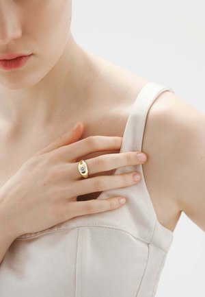 Mujer con camiseta blanca sin mangas tocándose el pecho, mostrando un anillo de oro con una gran piedra preciosa transparente en su dedo.