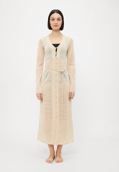 Lang, beige, strikket cardigan med ribbet tekstur, knaplukning og et fletbælte i taljen. Har lange ærmer og V-hals.