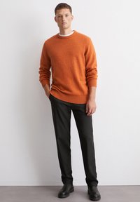 Oranges Strickpullover mit gerippten Bündchen und Saum, kombiniert mit schwarzen, gestreiften Hosen und schwarzen Stiefeletten. Glatte Textur, taillierte Passform.