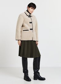 Veste en fausse peau de mouton beige avec bordures et boutons noirs, associée à une jupe plissée vert foncé et des bottes montantes noires.