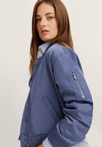 Helle blaue Bomberjacke aus glattem, glänzendem Stoff. Verfügt über einen gerippten Kragen und Bündchen, Seitentaschen und eine mit Reißverschluss verschließbare Ärmeltasche.