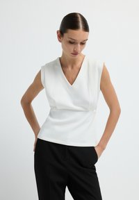 Top bianco senza maniche con scollatura a V e texture arricciata in vita, abbinato a pantaloni neri. Tessuto liscio e design strutturato.