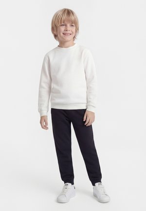 Lächelnder Junge, der steht und ein weißes Sweatshirt mit langen Ärmeln, schwarze Hose und weiße Turnschuhe vor weißem Hintergrund trägt.