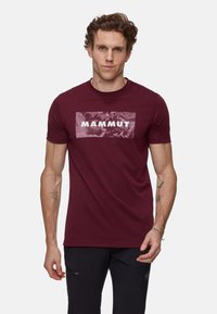 Burgundowa koszulka z krótkim rękawem wykonana z bawełny, z dużym, teksturowanym logo "MAMMUT" na przodzie na tle o nierównomiernym kolorze.