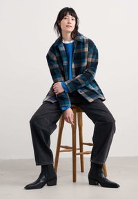 Mlad človek sedi na lesenem stolu, oblečen v modro karirasto jakno, modro pulover, črne široke kavbojke in črne gležnjarje.