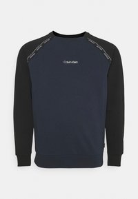 Sudadera azul marino con mangas negras, cuello redondo y logo de Calvin Klein en el pecho. Presenta una marca tonal a lo largo de las costuras de los hombros. Tela suave.