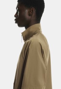 Braunes Kurzarmhemd mit hohem Kragen, lockerer Passform und glatter Textur. Enthält eine Kordelzugdetails in der Nähe des Kragens.