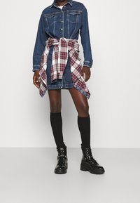 Jeanshemdkleid in Dunkelblau, mit Knopfleiste, zwei Brusttaschen und einem karierten Flanellhemd, das um die Taille gebunden ist. Schwarze kniehohe Socken und Stiefel.