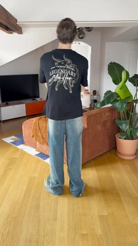 Juoda marškinėliai su baltu grafiniu dizainu nugaros dalyje, mėlynos plačios džinsinės kelnės, stovintys prieš sofą ir patalpų augalai šviesiame kambaryje.