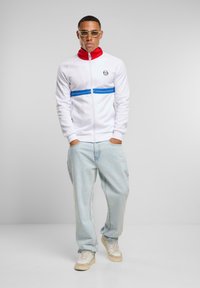 Giacca bianca con zip e colletto rosso, abbinata a jeans larghi blu chiaro e sneakers beidge. Occhiali trasparenti indossati.