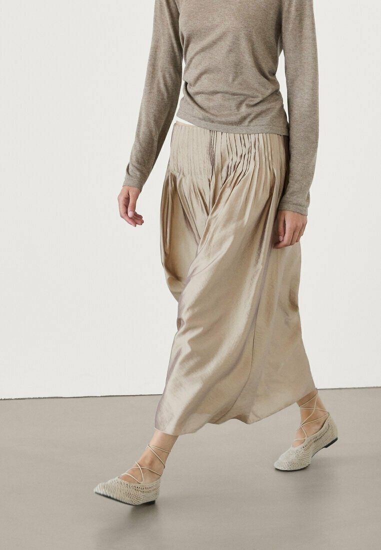 Personne portant une longue jupe en satin beige, un haut taupe à manches longues et des ballerines légères tissées avec des liens à la cheville, marchant sur un sol lisse.