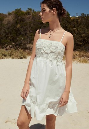 Robe d'été blanche avec un corsage en crochet ornée de détails floraux, des fines bretelles et un ourlet volanté. Portée sur un fond de sable.
