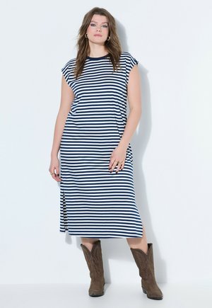 Kobieta w granatowo-białej sukience midi w paski z bocznymi rozporkami i brązowymi kowbojkami, stojąca na jednolitym białym tle.