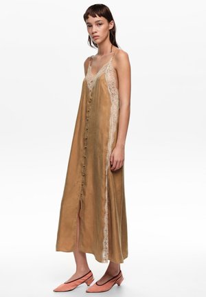 Joven mujer con un vestido de satén marrón tipo slip, con botones al frente y ribete de encaje crema, combinado con zapatos slingback coral de tacón bajo.