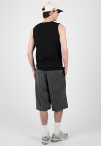 Homme portant un haut noir sans manches, un short gris à fines rayures jusqu’aux genoux, des chaussettes blanches, des baskets grises et une casquette beige, debout, dos tourné.