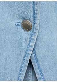 Světle modrá denimová tkanina s viditelným stříbrným kovovým knoflíkem a detaily šití. Zaměření na knoflík a texturu tkaniny.