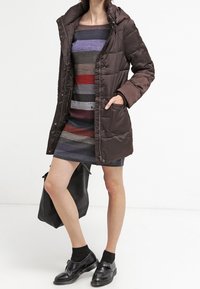 Doudoune marron avec capuche, dotée d'une texture lisse et de fermetures à bouton pression, portée sur une robe rayée multicolore. Sac et chaussures noirs.