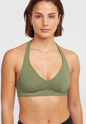 Young woman with short brown hair wearing an olive green halter sports bra and small gold hoop earrings against a plain background.