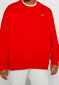 Czerwona bluza z bawełny, z długimi rękawami i okrągłym dekoltem. Zawiera małe logo na piersi oraz luźny krój.