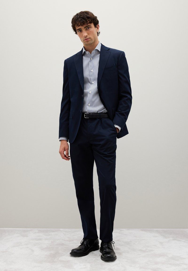 Giovane uomo in piedi con abito blu navy, camicia a righe con bottoni, cintura nera e scarpe eleganti nere davanti a uno sfondo chiaro uniforme.