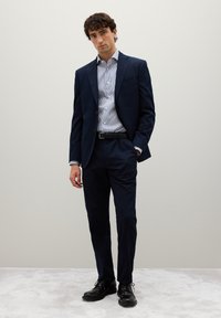 Mann steht mit einer Hand in der Tasche, trägt einen navy Anzug, ein gestreiftes Hemd, einen schwarzen Gürtel und schwarze Gesellschaftsschuhe vor einem schlichten Hintergrund.