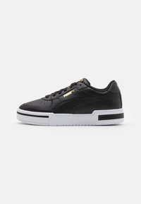 Zapatilla de cuero negra con una parte superior texturizada, suela de goma blanca, rayas en blanco y negro, y acentos de logo dorado en el lateral y la lengüeta.