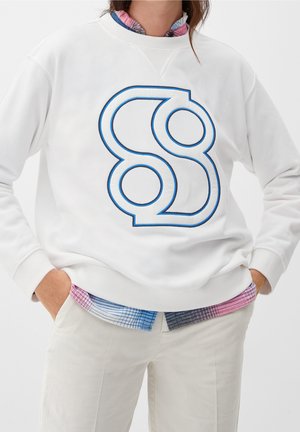 Person iført hvid sweatshirt med stor blå-omrids abstrakt grafik, lagdelt over pink og blå ternet skjorte, og hvide bukser, hænderne i lommerne.