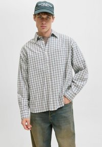 Młody mężczyzna noszący czarną czapkę, jasną koszulę w kratę na guziki, brązowy t-shirt pod spodem i wyblakłe zielone dżinsy, stojący na jednolitym tle.