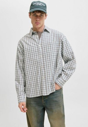 Young man wearing a black cap, light plaid button-up shirt, brown t-shirt underneath, and faded green jeans, standing against a plain background.