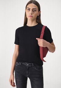 Kvinne i svart kortermet skjorte og mørke jeans som holder en liten rød skulderveske mot en enkel lys bakgrunn.