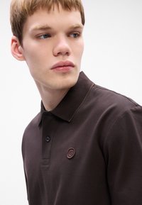 Mladý muž s krátkymi blond vlasmi, ktorý má na sebe tmavohnedé polo tričko s golierom a malým vyšitým logom na hrudi, pozerá sa na stranu.