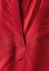 Tessuto rosso con una consistenza liscia presenta un profondo scollo a V e un dettaglio a punta. I punti di cucitura sono visibili lungo i bordi.